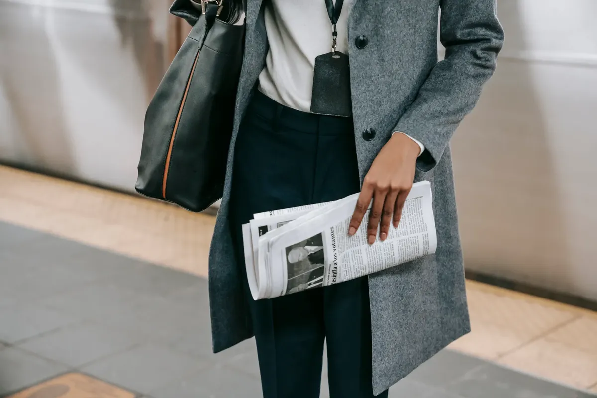 From above of crop anonymous female employee in elegant outfit and name tag standing on platform of metro station with newspaper in hand
